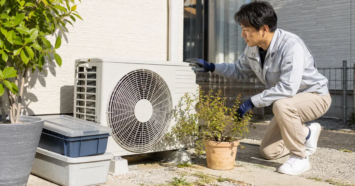 屋外でエアコンの室外機周辺に物が置かれていないか風通しを確認している男性の様子